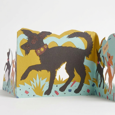
                    
                      Puppy Parade die-cut card, Mark Hearld illustration. Dark brown dog with red-brown collar, layered yellow & blue scene,…
                    
                  