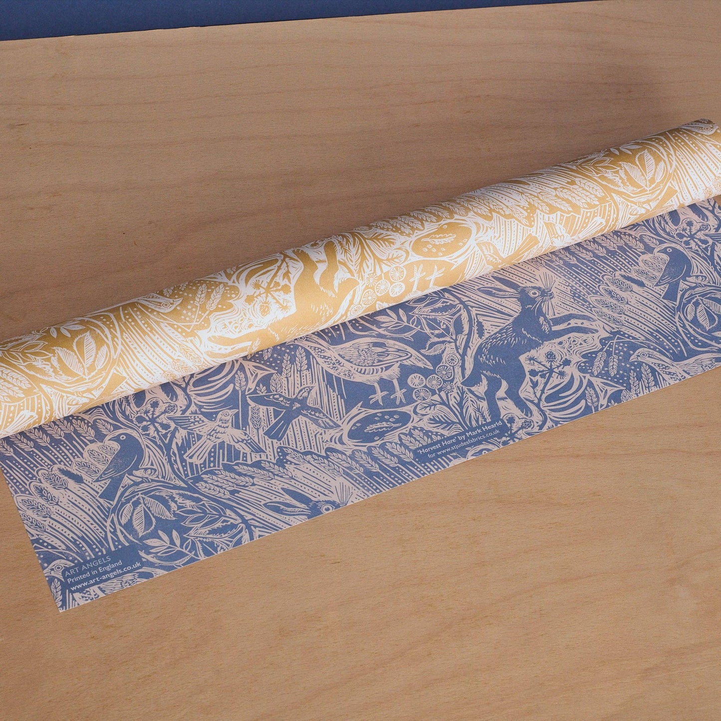Mark Hearld-Harvest Hare Double-Sided Wrapping Paper. Roll of patterned paper, one side is yellow and white, the other blue and white. Lying on a wooden surface.