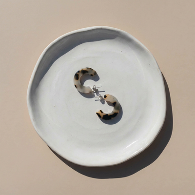 
                    
                      Ray ivory tortoise hoop earrings, cellulose acetate with stainless steel posts, flat lay pair on white ceramic dish.
                    
                  