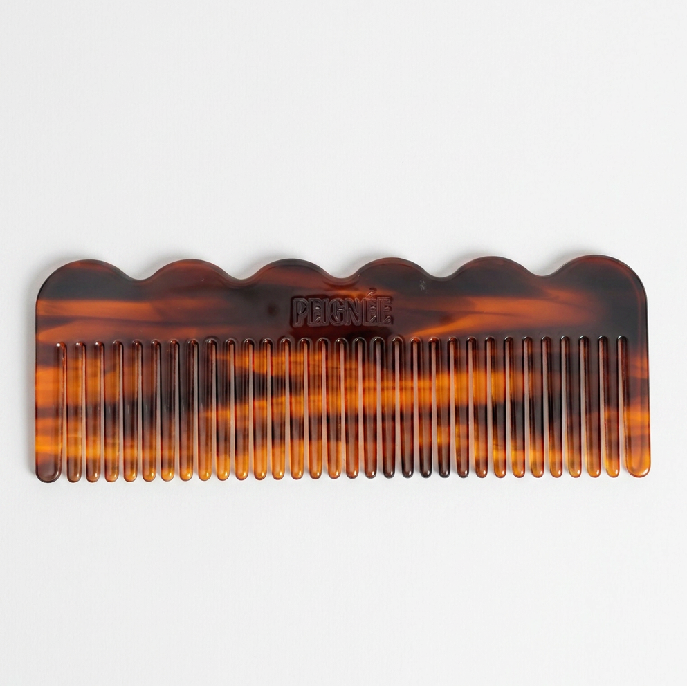 A tortoiseshell hair comb with a wavy scalloped edge, featuring a marbled amber, brown, and black pattern. The brand name 'PEIGNEE' is embossed on the top surface.