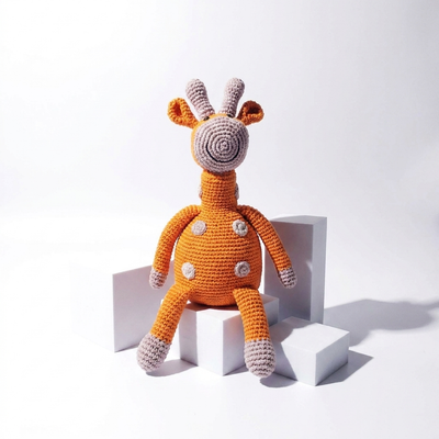 
                    
                      Pebble Curious Plush Giraffe, orange organic cotton crochet toy with grey details, sitting on white blocks, front view.
                    
                  