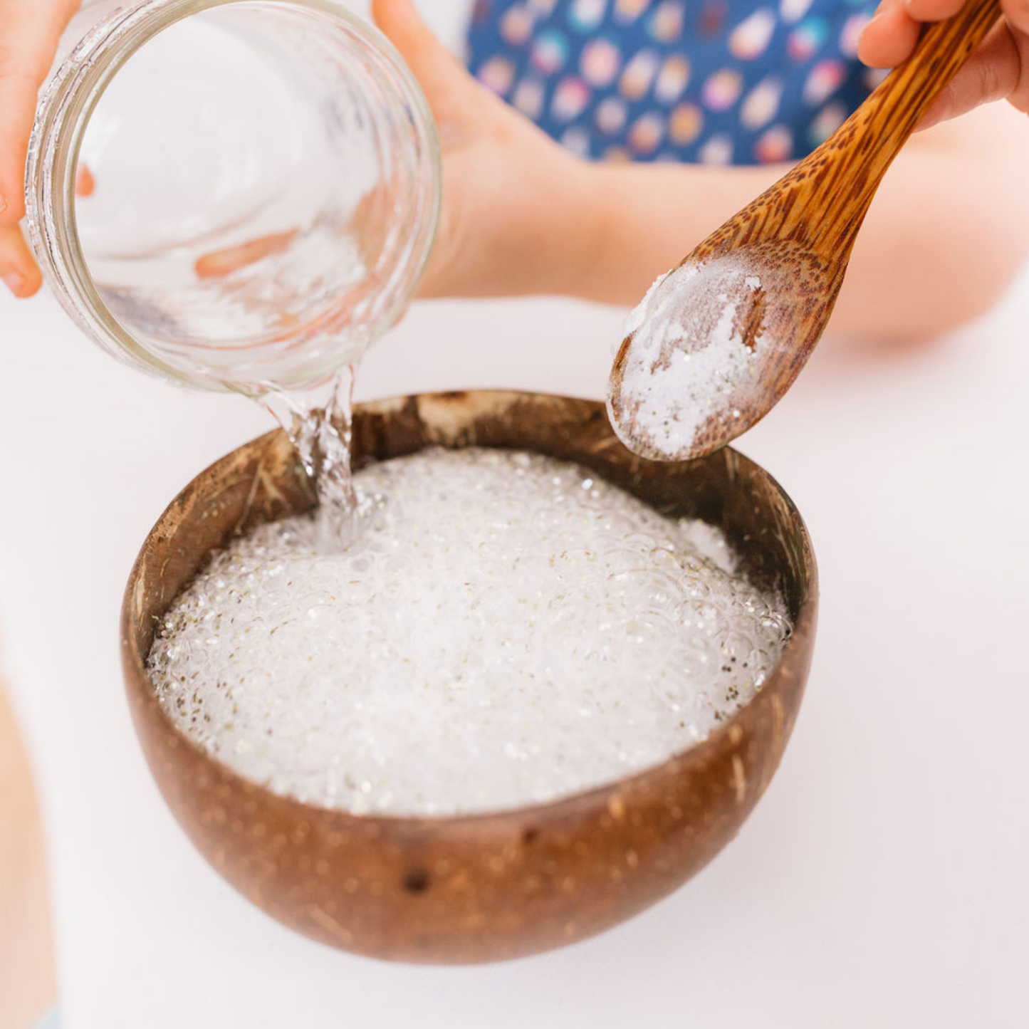 Wooden bowl with foamy liquid, person holding a spoon with more foam, and another person pouring from a jar.
