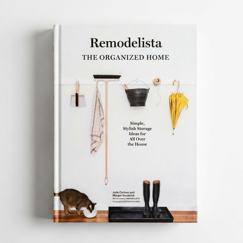 The Remodelista book, 'The Organized Home' by Julia Pincus and Megan Eversick, displayed upright against a clean white wall. The cover features minimalist home organization elements like a coat rack, bucket, and boots on a light background.