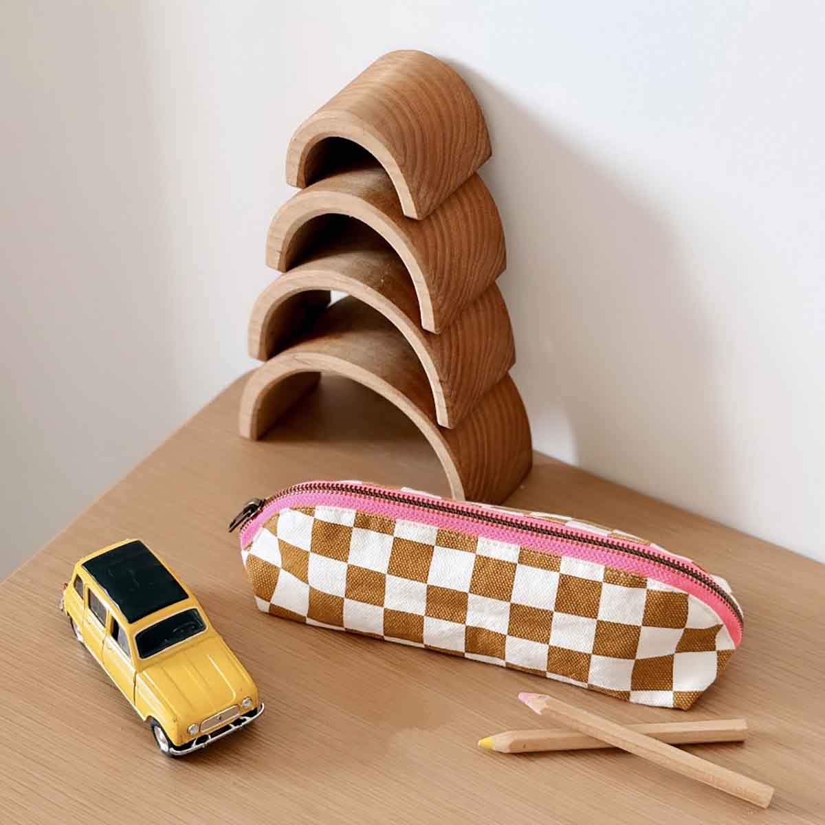 Checkered pencil case with pink zipper, yellow toy car, and wooden arches on a wooden surface.