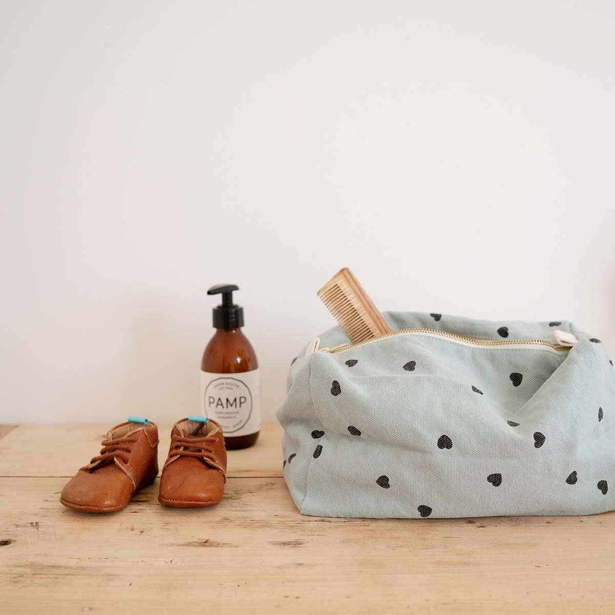 Rose in April: Vic Toiletry Bag (Blue Heart). Baby shoes, bottle, and bag on a wooden surface with a white background