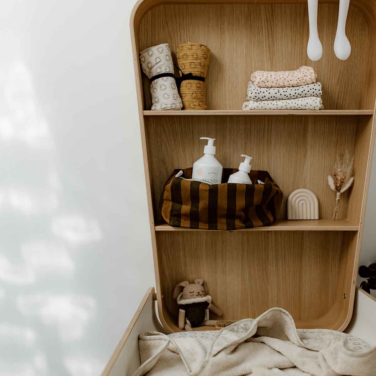 Rose in April: Vic Toiletry Bag (Cumin). Wooden shelf with various items, including baskets, bottles, and a toy, on a white wall.