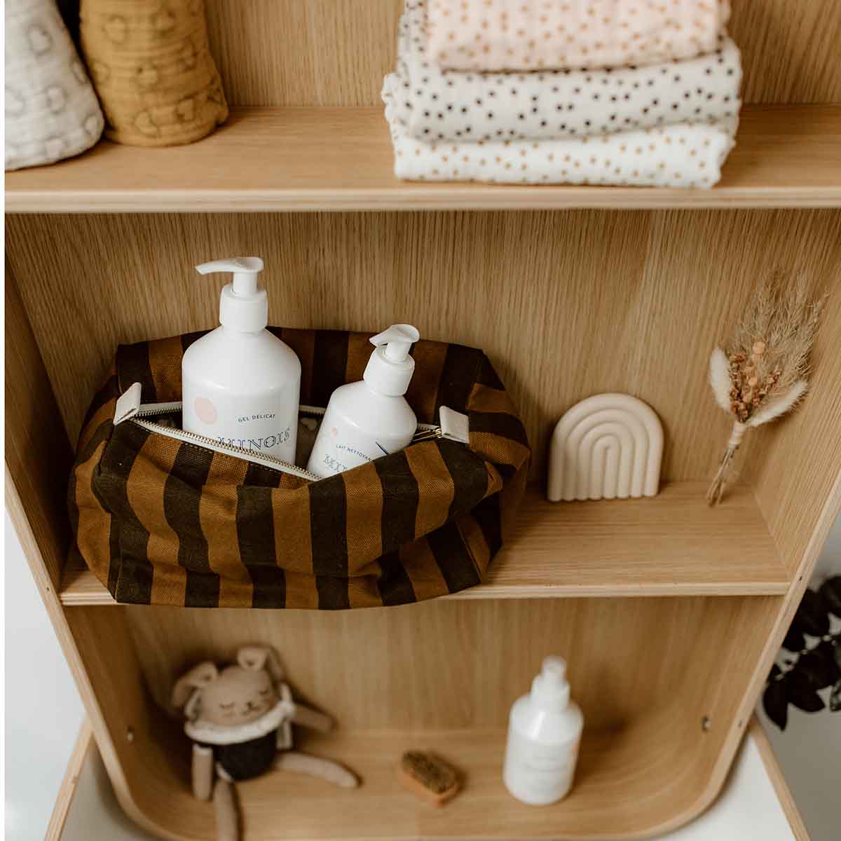 Rose in April: Vic Toiletry Bag (Cumin). Wooden shelf with various items, including bottles, a striped basket, and decorative objects.