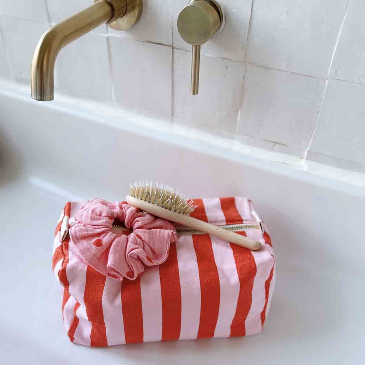 Rose in April: Vic Toiletry Bag (Tomato + Candy Pink). Red-and-white striped pouch with a scrunchie and a brush on a white sink.
