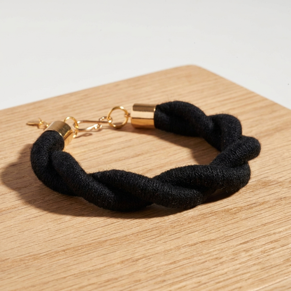Sasha Hemp Twist Bracelet, black fiber-wrapped cording with raw brass hardware, flat lay on a light wood surface.