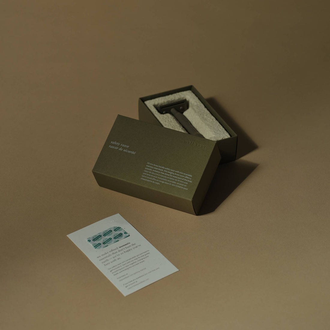Kelp brass safety razor in open green box, nestled on white towel, alongside stainless steel blades and info card.