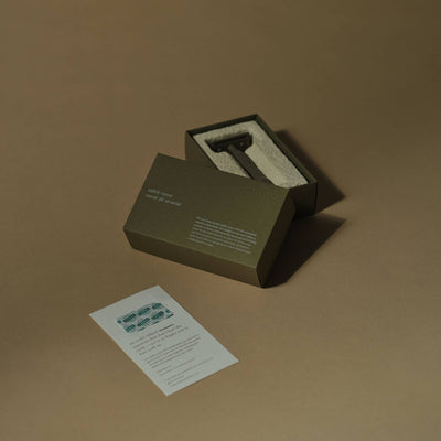 
                    
                      Kelp brass safety razor in open green box, nestled on white towel, alongside stainless steel blades and info card.
                    
                  