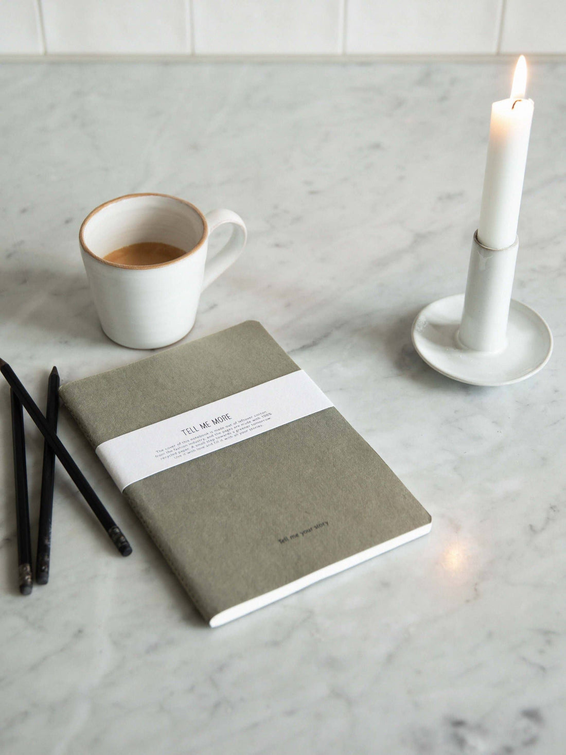 Tell Me More Recycled Notebook, green-grey cardboard cover with white band, angled flat lay on marble, next to pencils and…