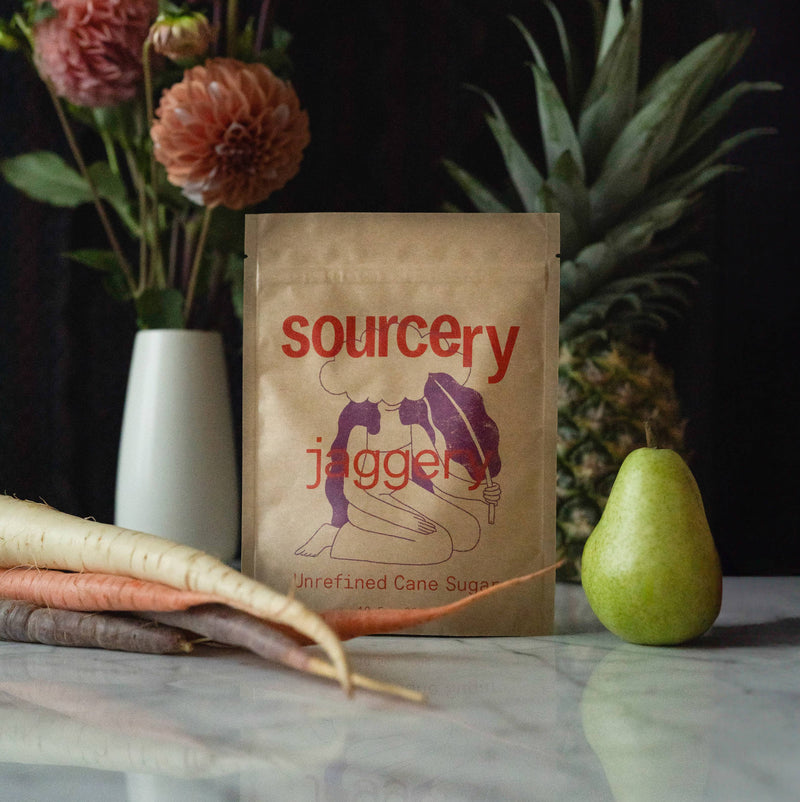 Sourcery Artisanal Jaggery sweetener, kraft bag with red text & purple illustration, front view on marble with fresh produce.