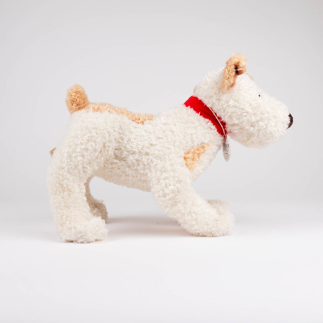 Eliot Stuffed Dog, cream plush with tan patches and red fabric collar, full side view, product tag visible.