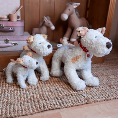 
                    
                      Trio of Eliot Stuffed Dogs in different sizes, white plush with tan patches and red collars, sitting on a jute rug.
                    
                  
