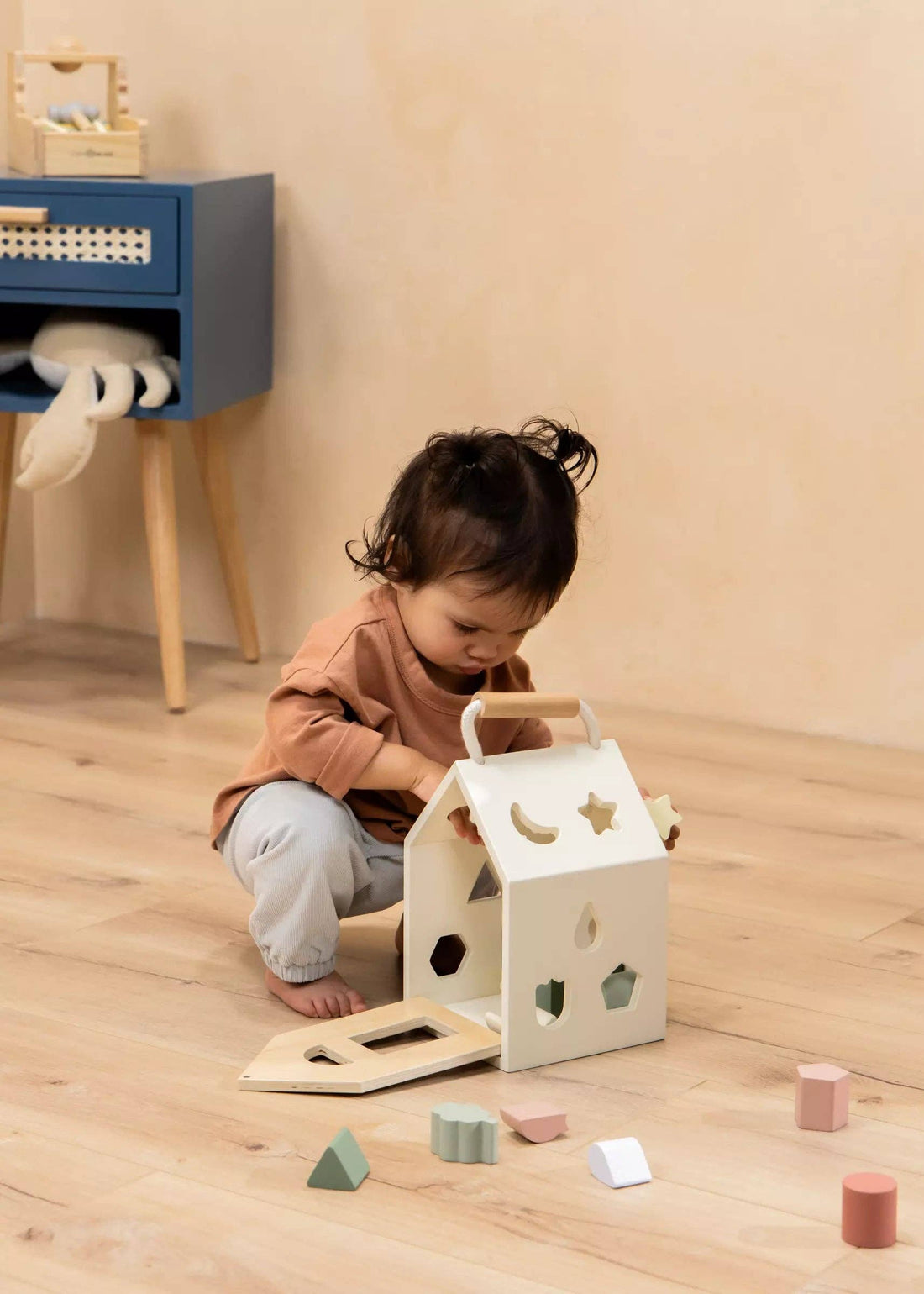 Wooden Shapes Sorting House, creamy white solid wood, child playing on floor with scattered pastel blocks