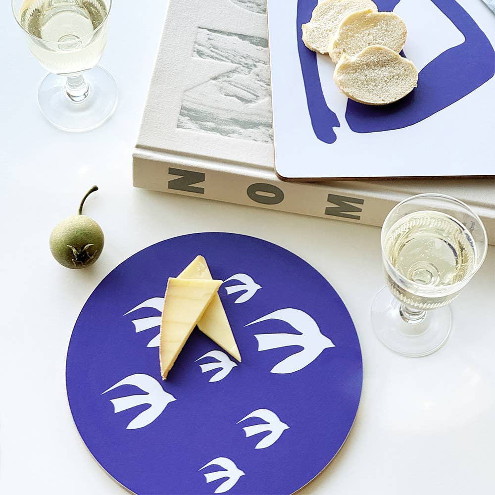 A styled tabletop scene showing a round blue tray with white bird motifs, topped with two slices of cheese, alongside a matching square plate with bread slices, books, and two wine glasses on a white surface.