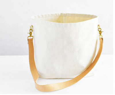 
                    
                      Everyday Tote Bag, natural cotton canvas, open top showing interior, tan leather strap with brass clips, angled view.
                    
                  