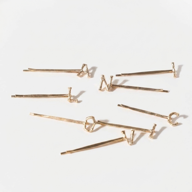 Peignee Paris Alphabet Letter gold-plated steel hair clips, featuring textured letters, scattered flat lay on white.