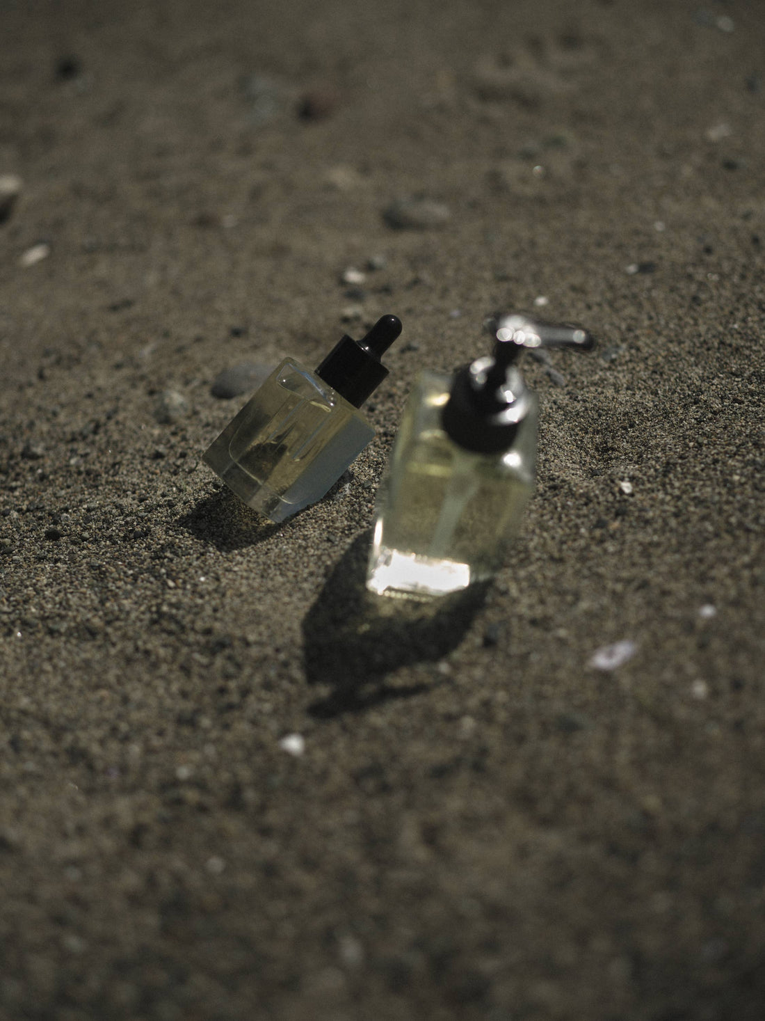 Shave Oil, two clear glass bottles with black caps on dark sand. One dropper bottle in focus, one pump dispenser blurred.