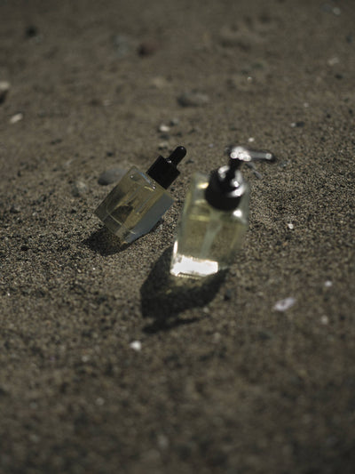 
                    
                      Shave Oil, two clear glass bottles with black caps on dark sand. One dropper bottle in focus, one pump dispenser blurred.
                    
                  