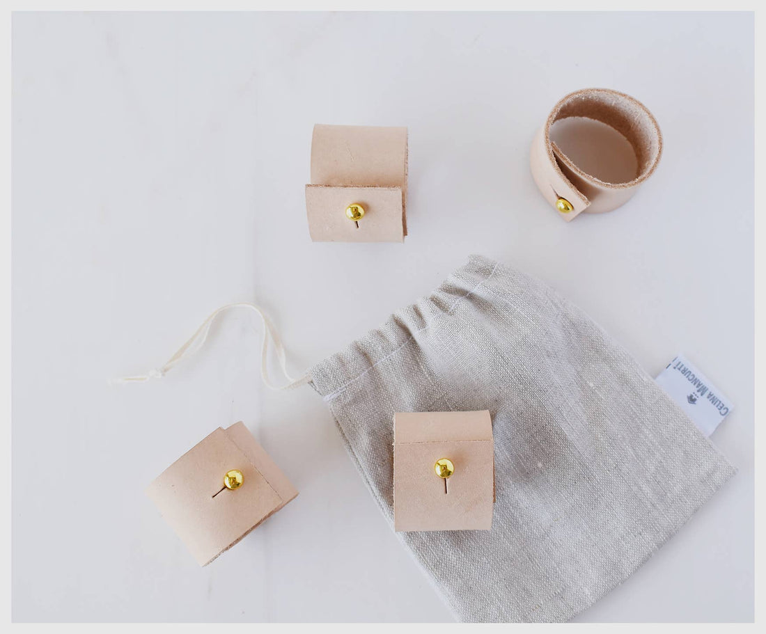 Tan leather Napkin Rings, set of three, with brass clips, alongside light grey linen storage bag, flat lay.