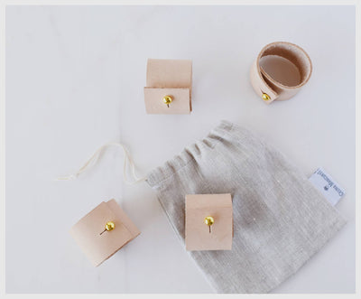 
                    
                      Tan leather Napkin Rings, set of three, with brass clips, alongside light grey linen storage bag, flat lay.
                    
                  