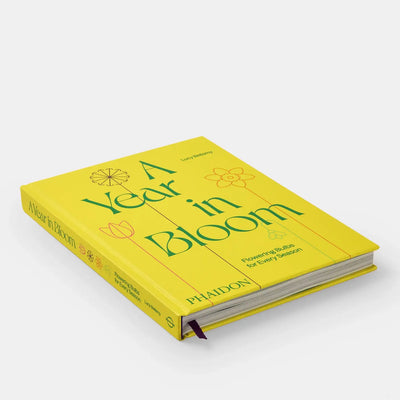 
                    
                      Phaidon A Year in Bloom hardcover book, bright yellow cover with green text, angled view showing spine and purple ribbon…
                    
                  