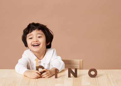 
                    
                      Happy child holds a two-tone beech and walnut 'B' Alphabet Play Block. Walnut 'I', 'N', 'O' blocks on light wood table.
                    
                  