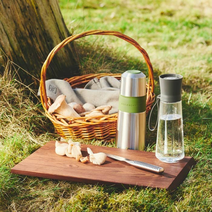 Rå Ash Wood Chopping Board, dark heat-treated ash, with foraged mushrooms and a knife on grass outdoors.