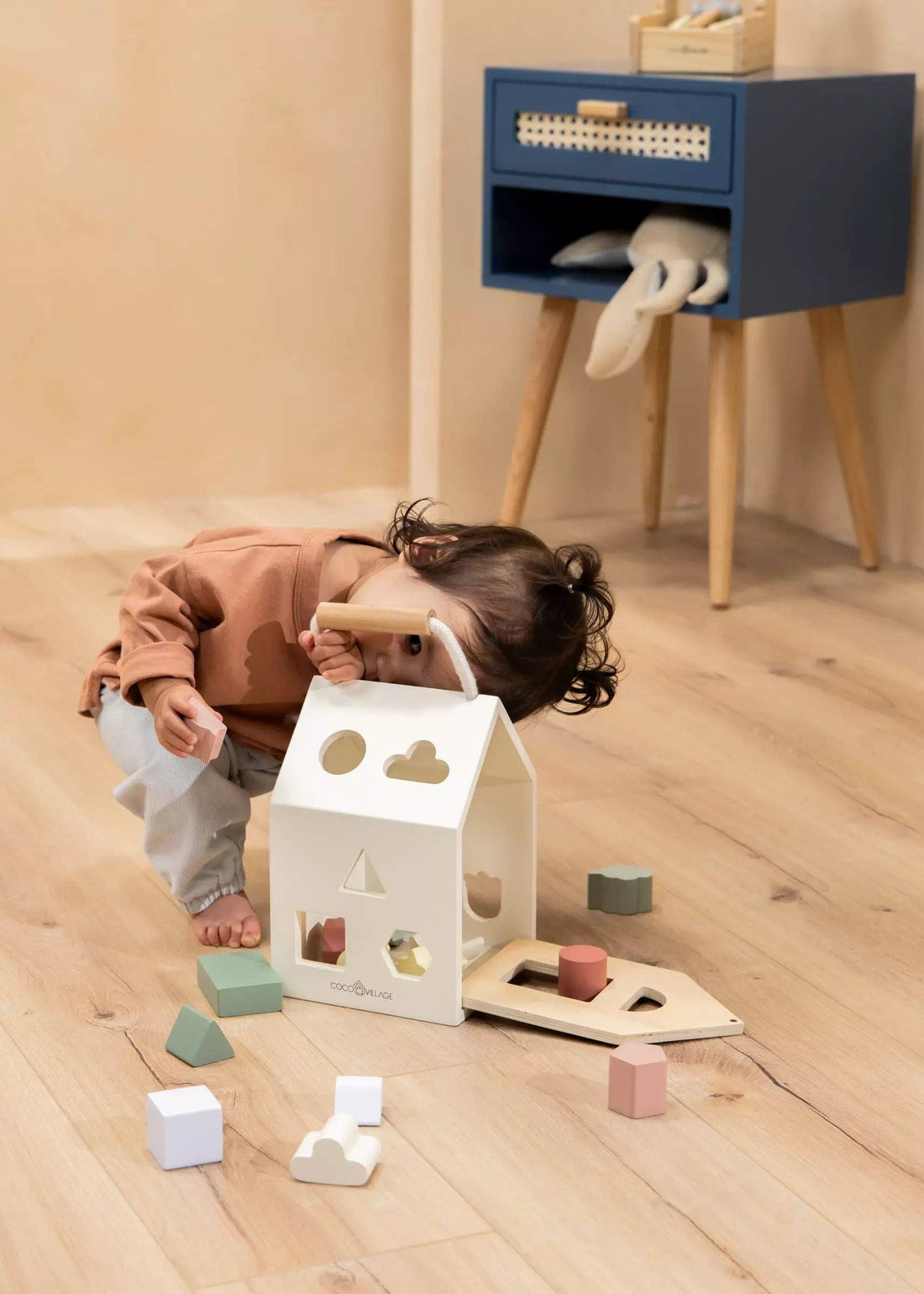 Wooden Shapes Sorting House, white solid wood, toddler peeking inside, pastel blocks on light wood floor.