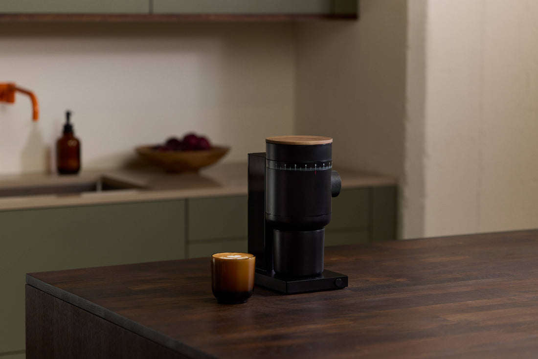 Opus 2 Conical Burr Grinder, black with natural wood lid, on dark wood kitchen island next to a latte.