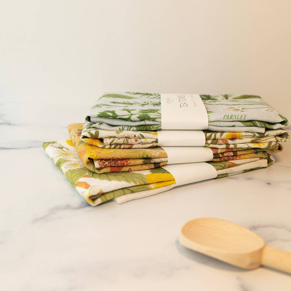 Stacked Herb Garden Linen Tea Towels, folded with paper bands, displaying diverse green and yellow botanical patterns.