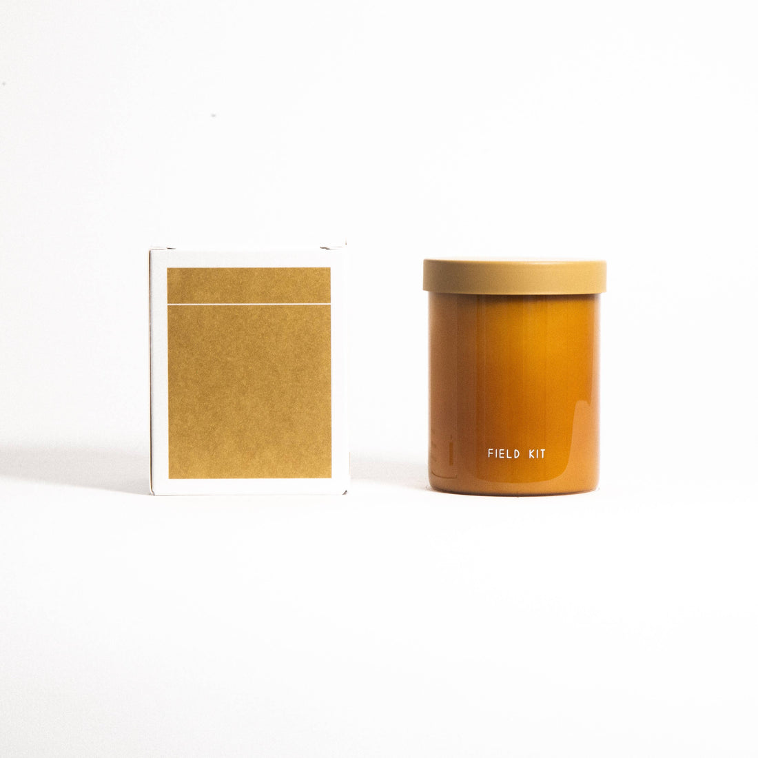 Field Kit Beekeeper Glass Candle, honey-amber glass jar with tan lid, shown side-by-side with its white and gold packaging…