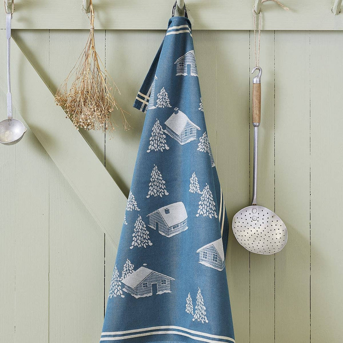 Torchon Jacquard Coeur Vallée tea towel, blue 100% cotton with white cabins & trees jacquard pattern, hanging against wood…
