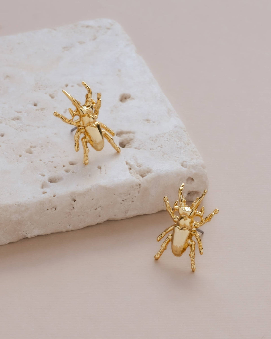 Mayana Designs Gold Beetle Stud Earrings, 24K gold-plated, angled flat lay pair on textured stone.