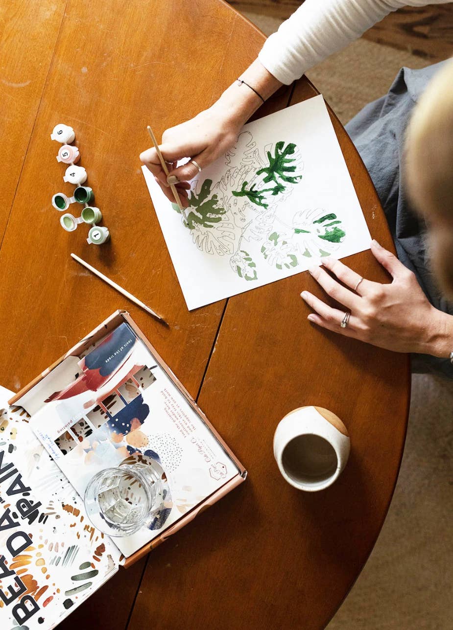 Peony Blossoms paint-by-numbers kit, person painting green leaves with acrylics, paint pots, brush, and kit box on table,…