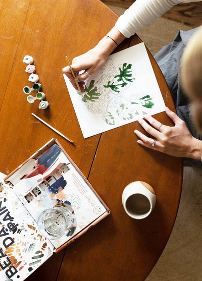 
                    
                      Peony Blossoms paint-by-numbers kit, person painting green leaves with acrylics, paint pots, brush, and kit box on table,…
                    
                  