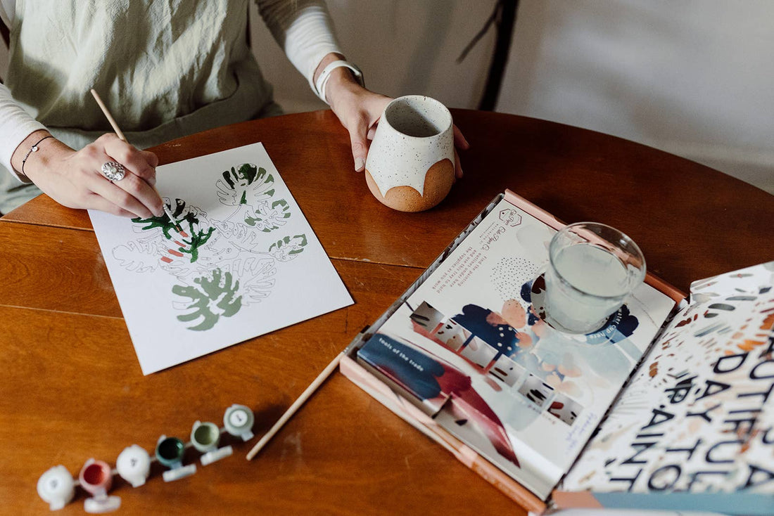 Peony Blossoms Paint-by-Numbers Kit. Hands apply green acrylic paint to white paper design, with other paint pots and brush.