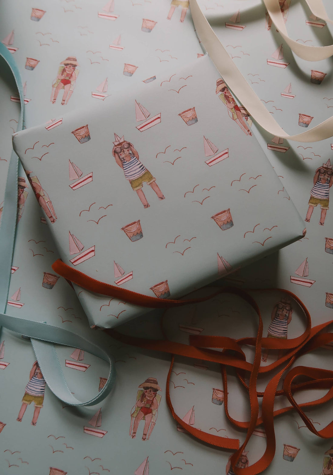 Summer Wrapping Paper, Agnese Grīnberga illustration, light blue beach scene, wrapped box with scattered ribbons.