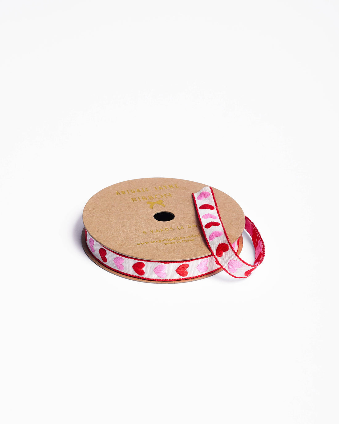 Hearts woven ribbon, white fabric with alternating pink and red embroidered hearts and red trim, close-up on spool.