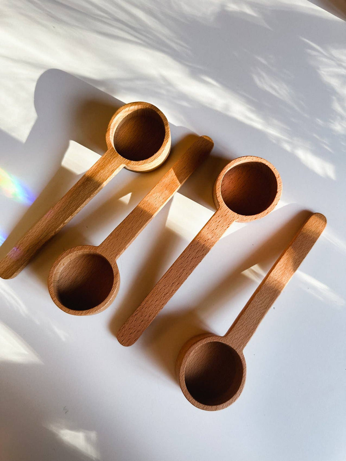 Wooden Tea Scoop, four unfinished beechwood scoops, flat lay, arranged diagonally with sunlight on white.