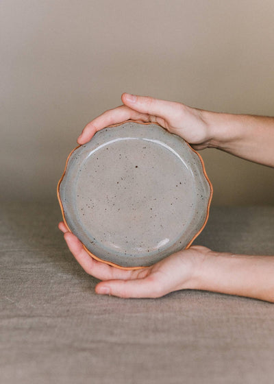
                    
                      Handcrafted Stoneware Plate, speckled gray, with wavy raw brown rim, held in hands for scale and detail.
                    
                  