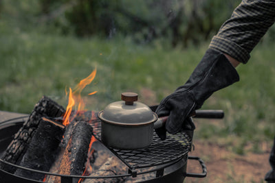 
                    
                      Enamel Saucepan with Walnut Handle, gray, with lid knob, cooking on campfire grill, held by a gloved hand.
                    
                  