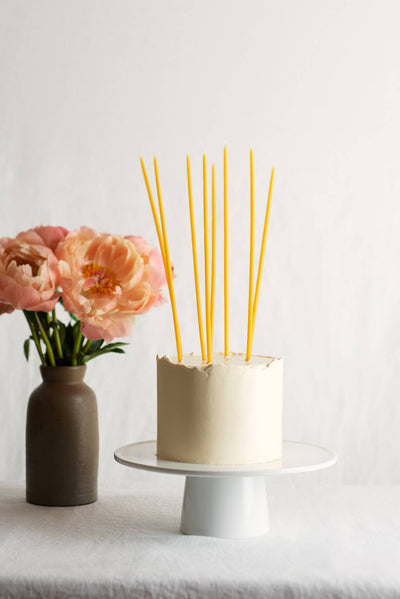 
                    
                      Multiple Fête Tall Beeswax Birthday Candles, natural yellow, standing in a white frosted cake, with pink peonies in a vase.
                    
                  