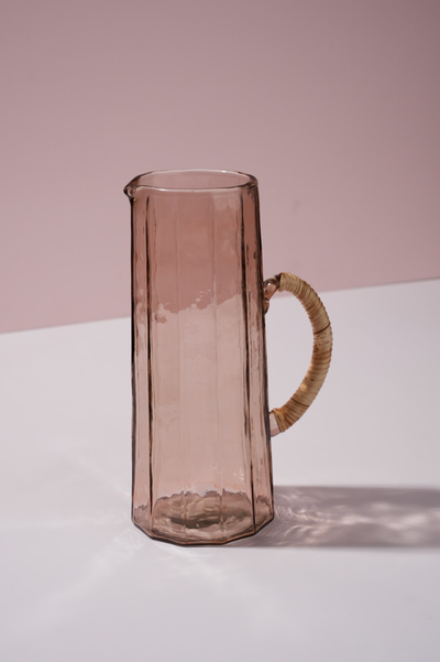 
                    
                      Hammered Glass Pitcher, rose-tinted handblown glass with natural woven rattan handle, full front view detail.
                    
                  