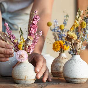 Hands arranging pink and yellow dried flowers in textured cream vases, a visual from 'Designing with Dried Flowers'.