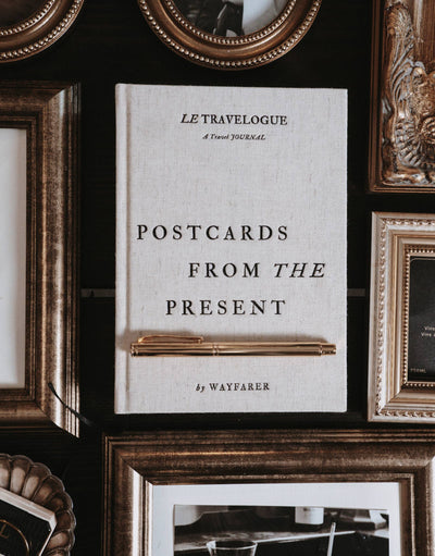
                    
                      Wayfarer Postcards from the Present Travel Journal, light linen cover, with a gold pen resting on it, flat lay among ornate…
                    
                  