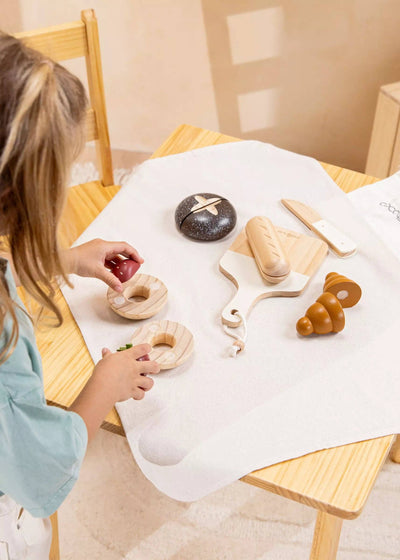 
                    
                      Wooden Bakery Set toys: child's hands playing with natural wood sliced doughnuts and a red berry half on table.
                    
                  