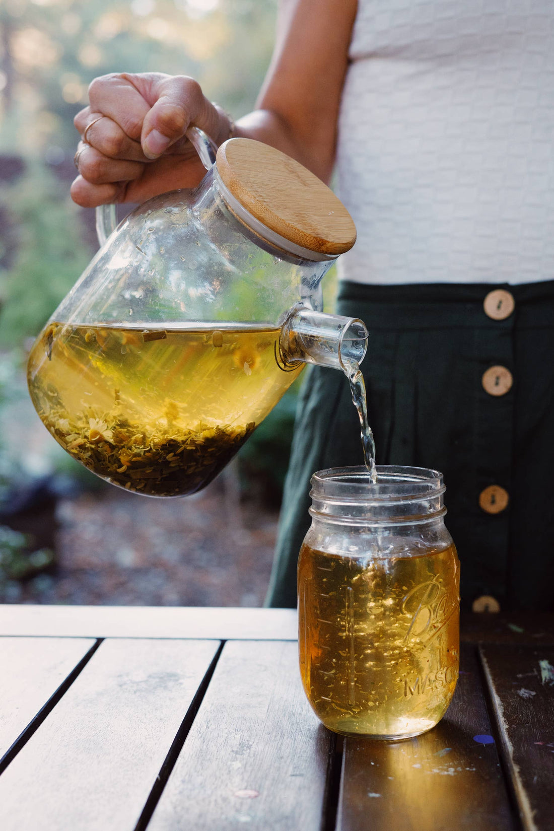 Heat-Resistant Glass Teapot, borosilicate glass & bamboo lid, hand pouring chamomile tea into glass jar outdoors.